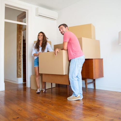 Positive young couple looking over their new apartment, while standing and leaning on cardboard boxes and furniture indoors. Full length. New home or property buying concept