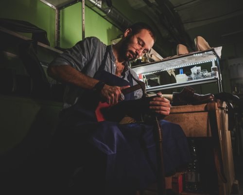 Young cobbler in glasses is working on shoe sole at his dark workplace.