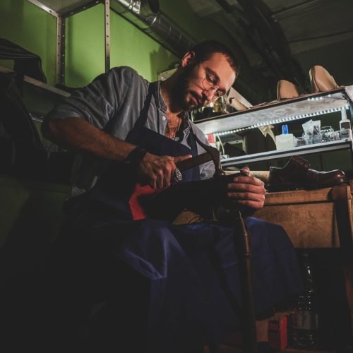 Young cobbler in glasses is working on shoe sole at his dark workplace.